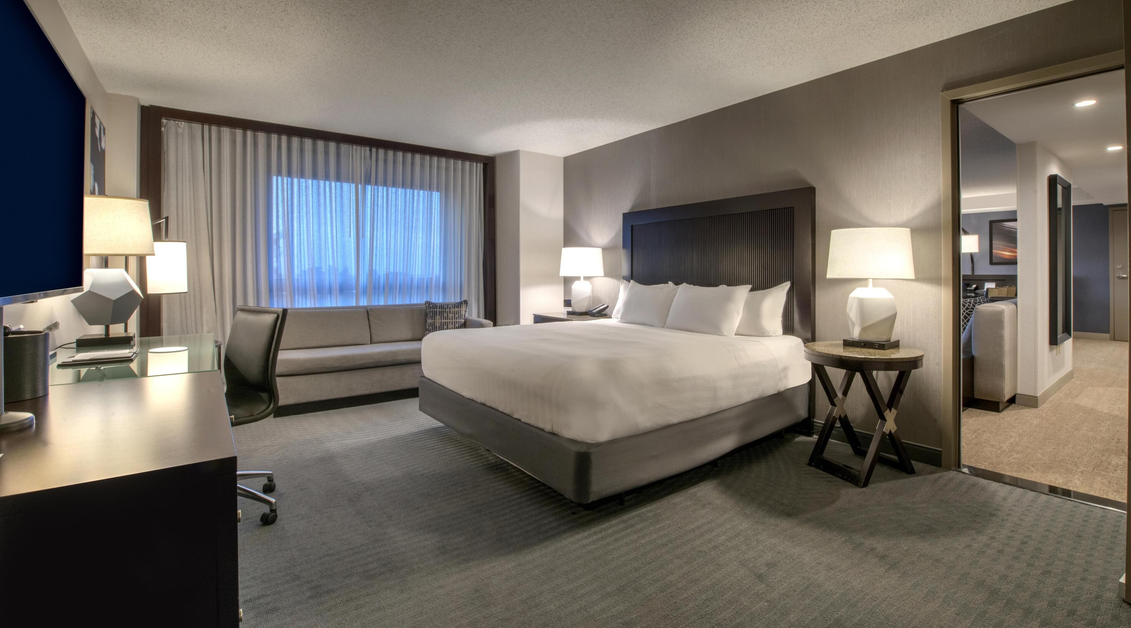 A modern hotel room with a large bed, two bedside tables with lamps, a desk with a chair, a sofa, and a large window with curtains.