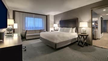 A modern hotel room with a large bed, two bedside tables with lamps, a desk with a chair, a sofa, and a large window with curtains.