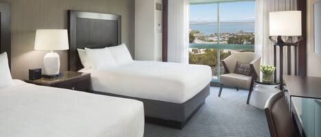 Ein Hotelzimmer mit zwei Betten, einem Schreibtisch, einem Stuhl und einem großen Fenster mit Blick auf die Stadt und das Wasser.