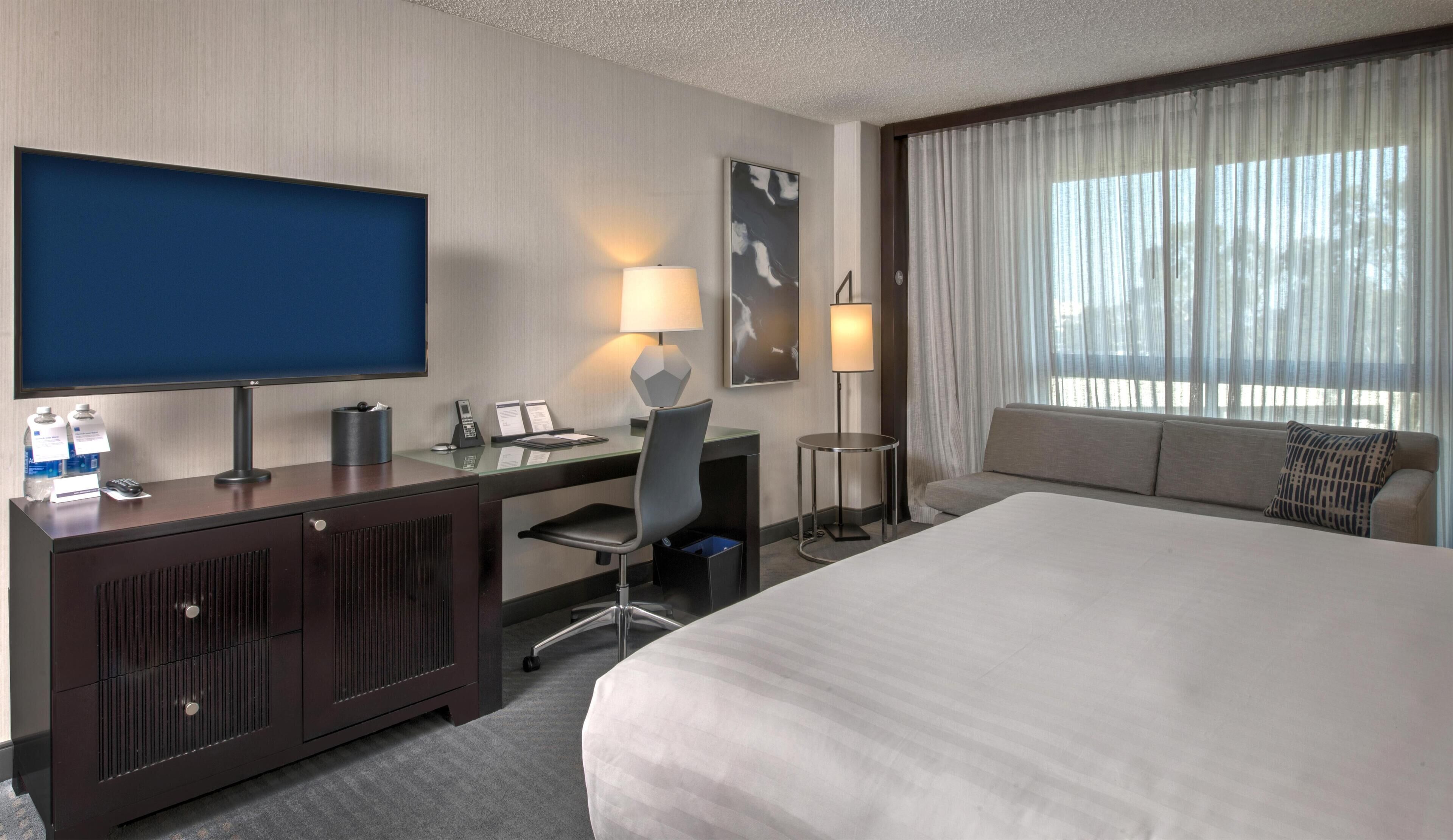 A hotel room with a large bed, a desk with a computer, a flat-screen TV, two lamps, a sofa, and a window with curtains.