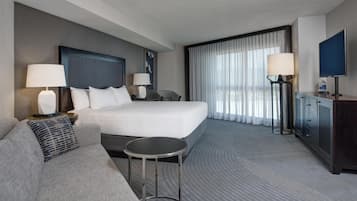 A hotel room with a large bed, a sofa, a small table, a TV, and a desk with a lamp.