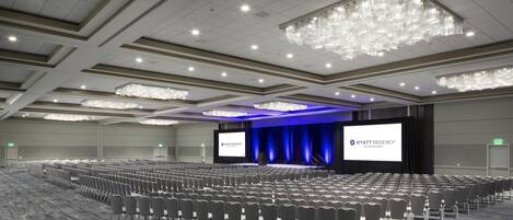 Uma grande sala de conferências com fileiras de cadeiras, um palco com uma tela exibindo 'HYATT REGENCY' e um lustre.