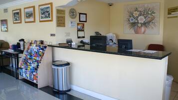 A hotel reception area with a counter, a trash bin, framed pictures on the wall, and a flower painting.