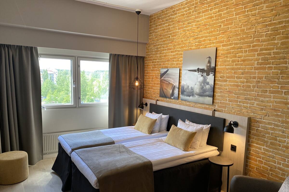 A hotel room with a large bed, two bedside tables, a view of greenery outside, and a brick wall.