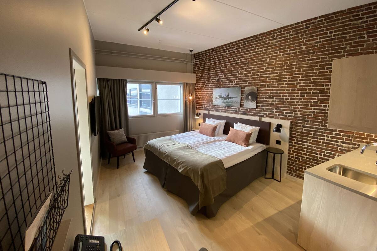 A modern hotel room with a brick accent wall, a large bed with multiple pillows, a desk with a sink, and a chair.