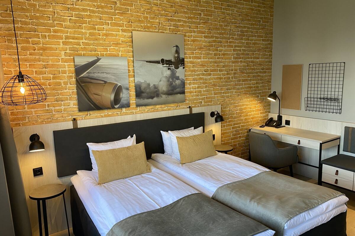 A hotel room with a brick wall, a bed with white linens, two bedside tables with lamps, a desk with a chair, and artwork on the wall.
