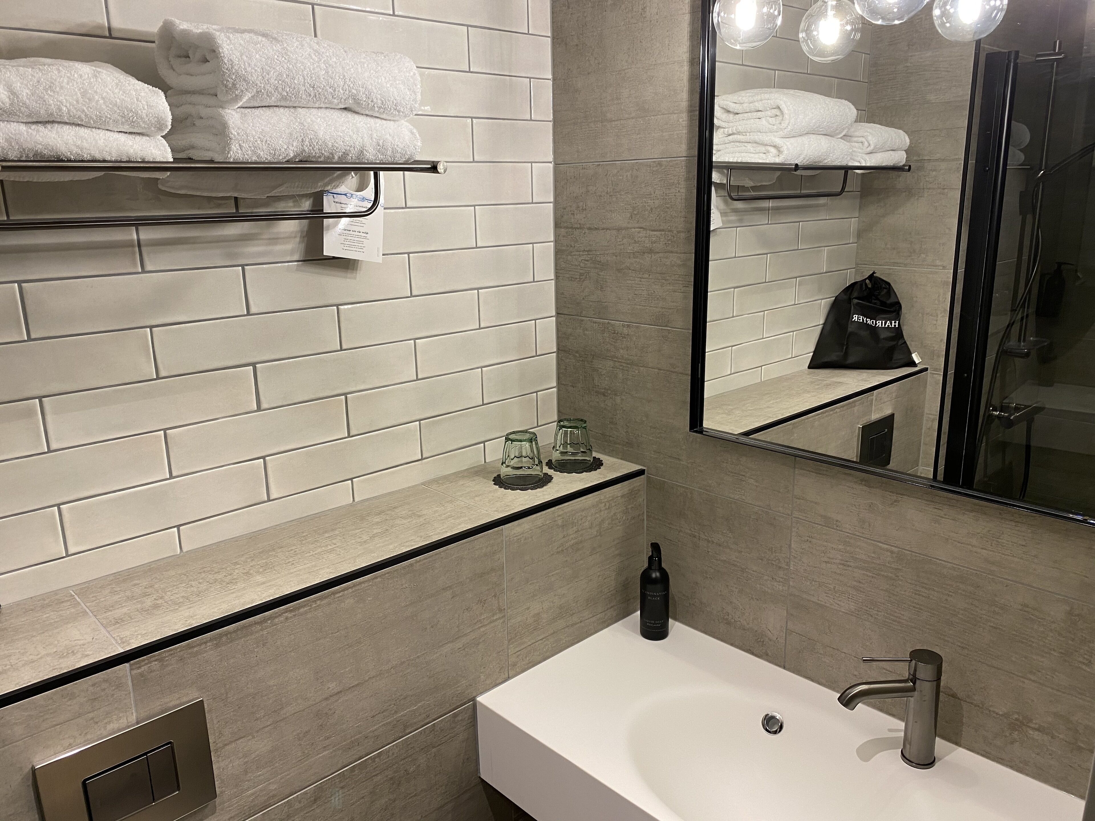 A modern bathroom with a white sink, a glass shower enclosure, and a mirror reflecting a towel rack and a bag.