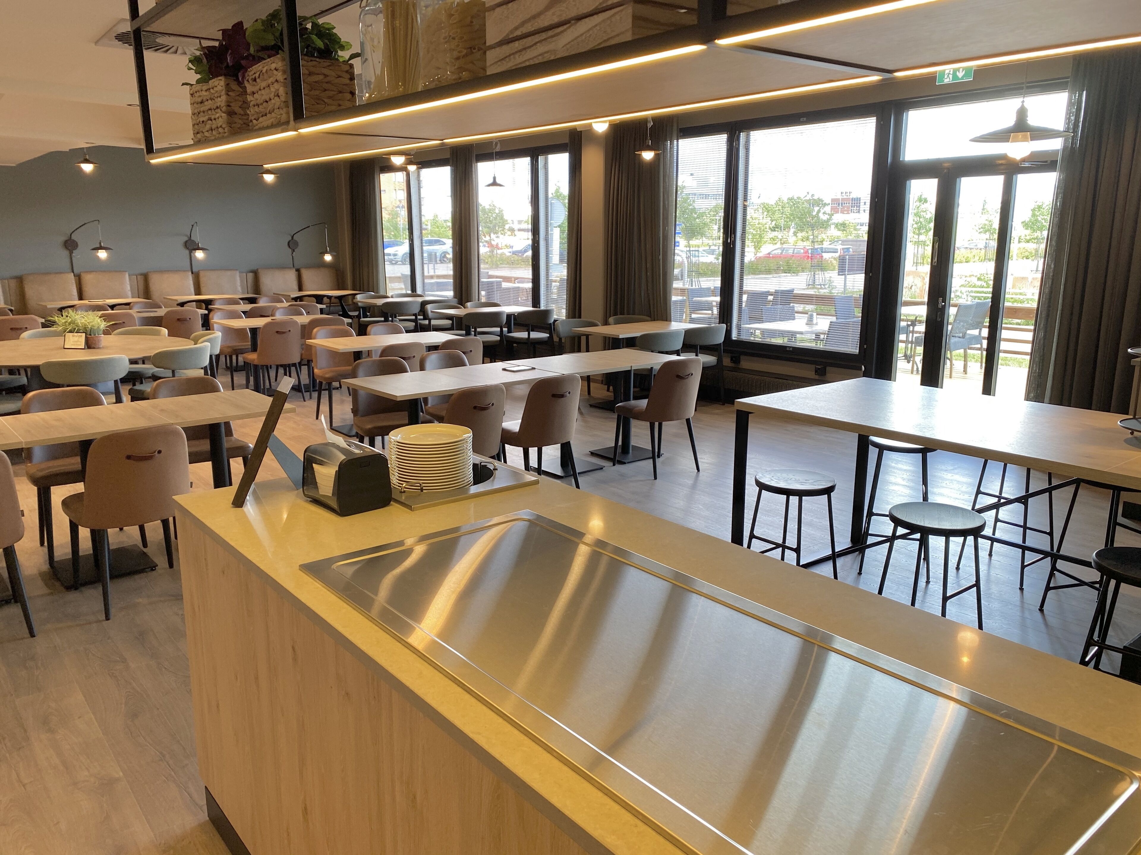 A modern dining area with multiple tables and chairs, a central stainless steel sink, and large windows providing natural light.
