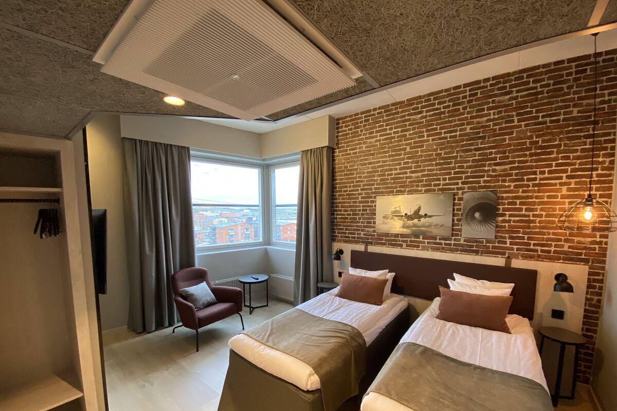 A hotel room with two beds, a brick wall, a large window, a red armchair, and a small table.