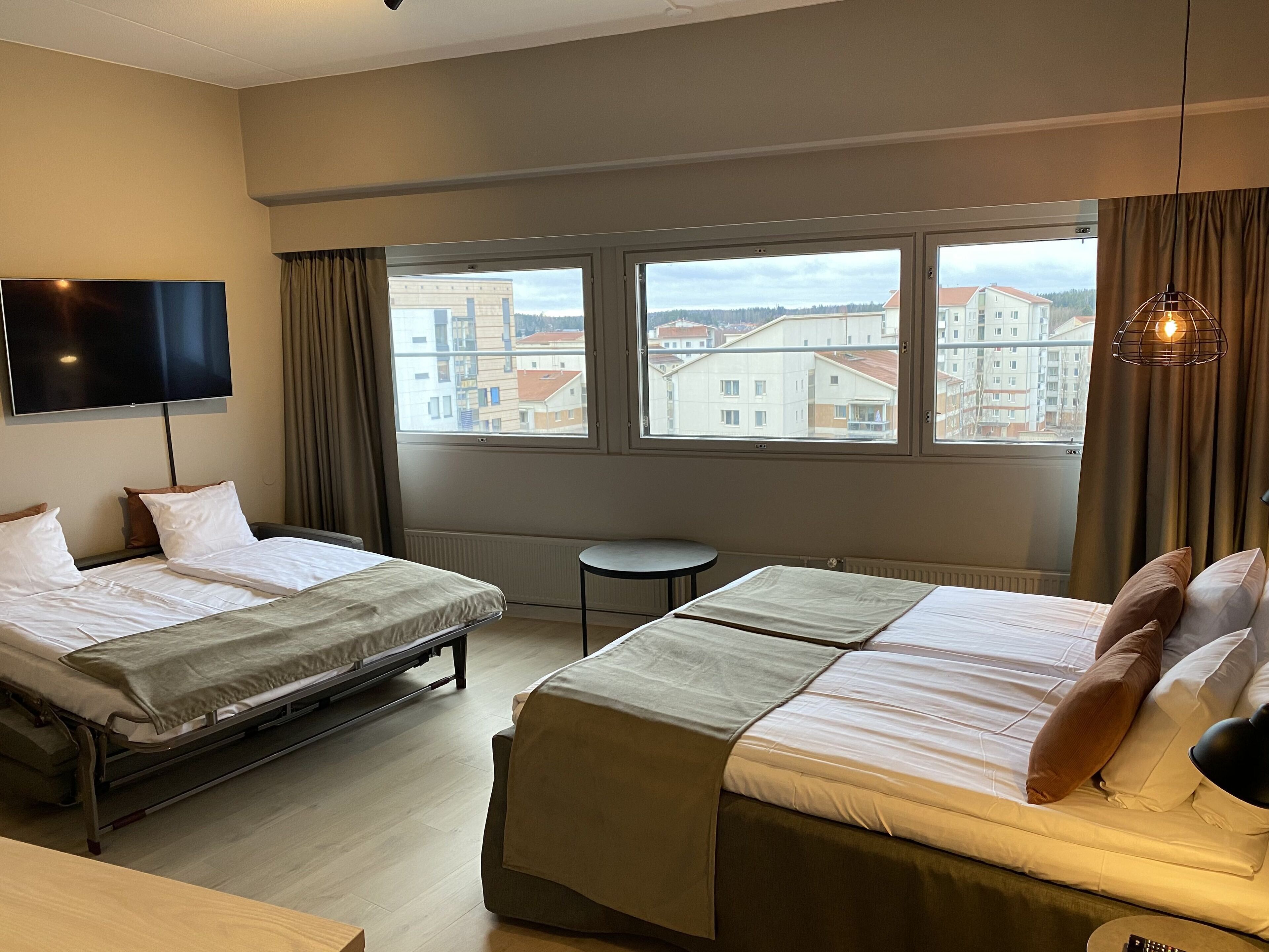 A hotel room with two beds, a TV, a small table, and a view of buildings through large windows.