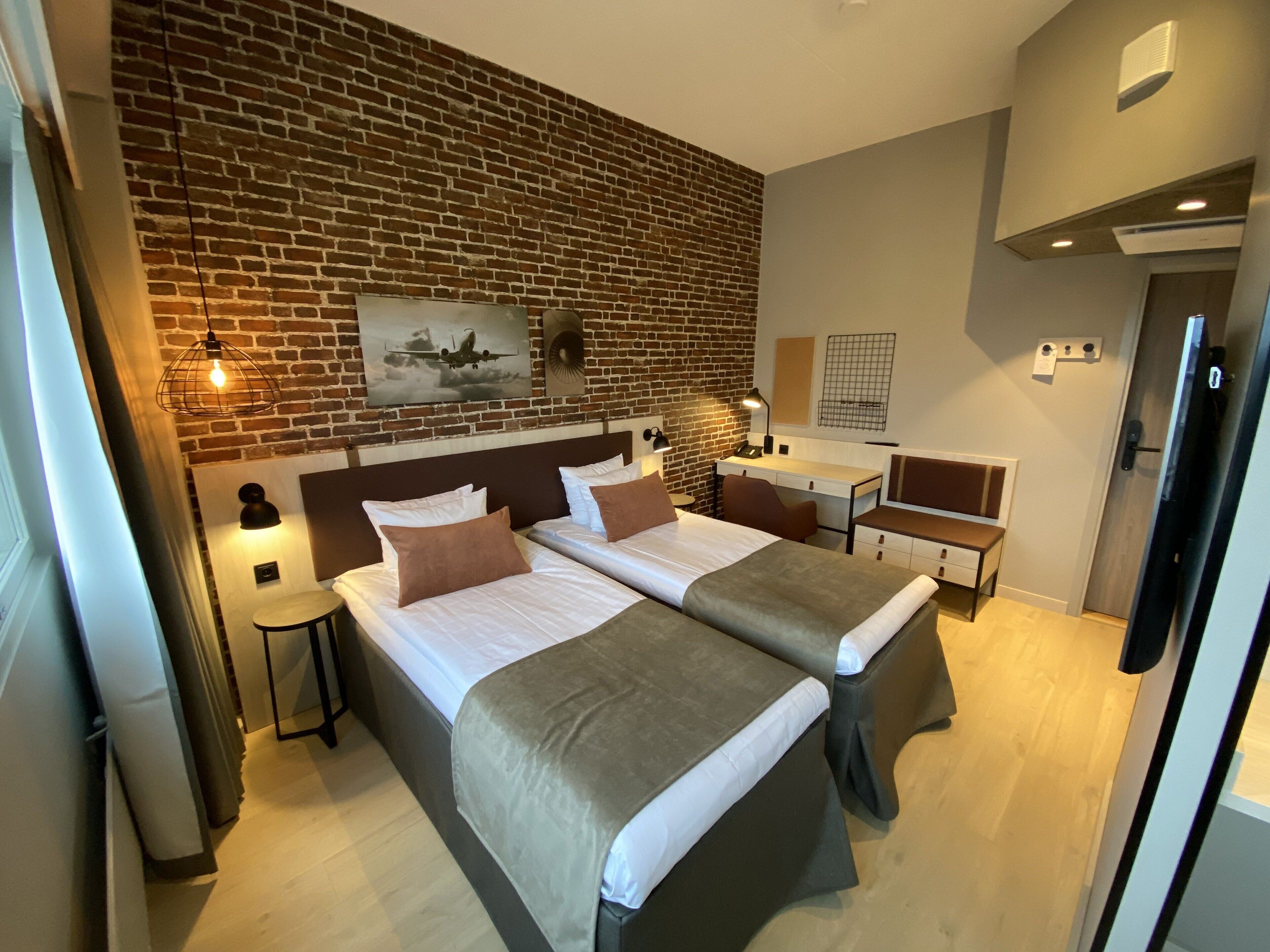 A hotel room with two beds, a desk, a chair, a small table, and a brick wall.