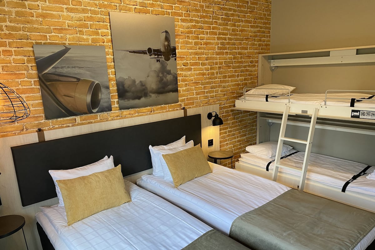 A hotel room with two single beds, a bunk bed, a brick wall, and two framed pictures.