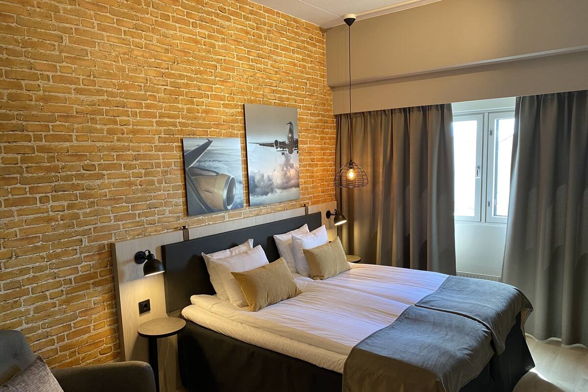 A hotel room with a brick wall, a large bed with multiple pillows, a small round table, a sofa, and a window with curtains.