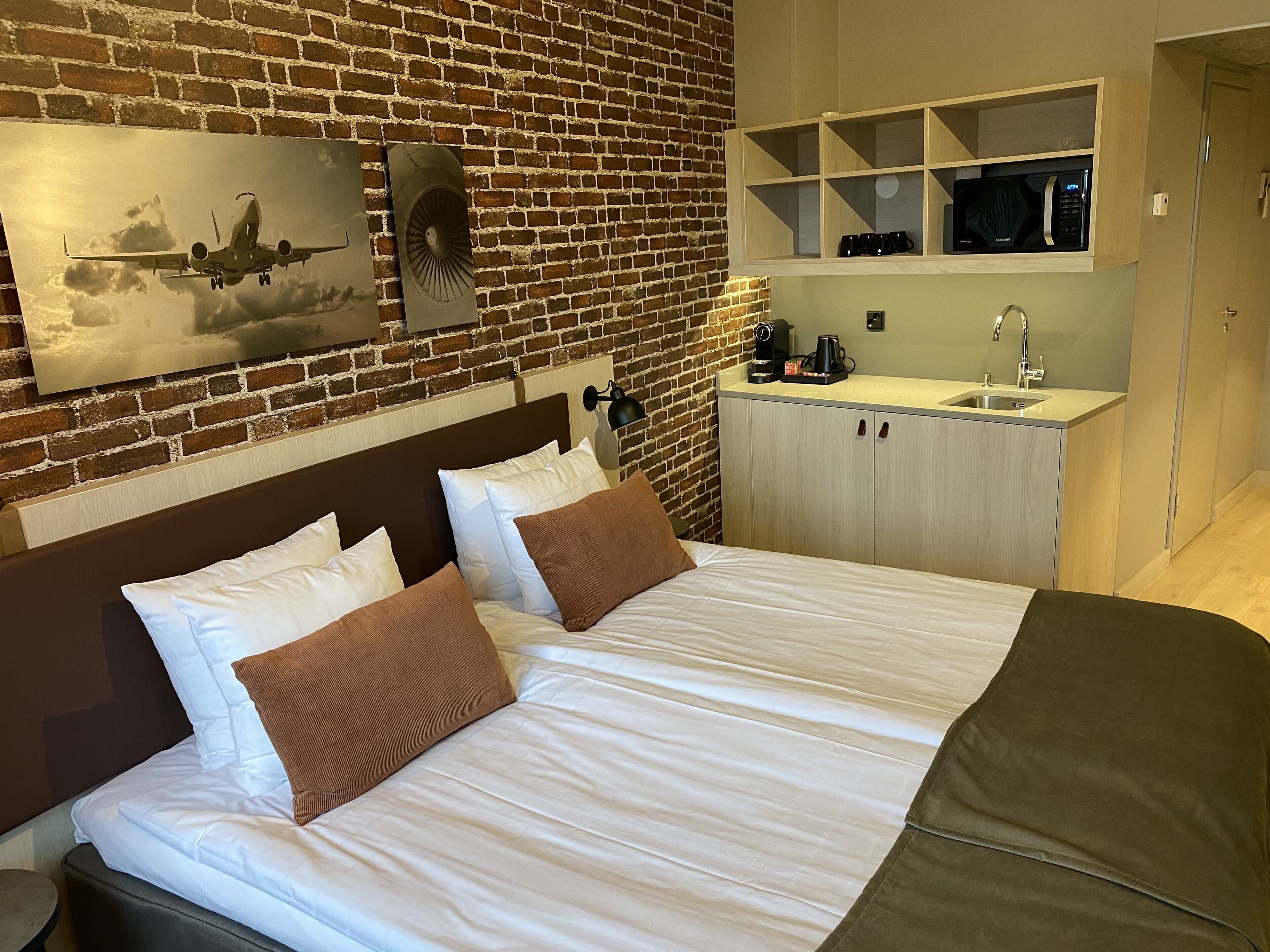 Une chambre d’hôtel avec un mur en briques, un lit avec une literie blanche et marron, une petite kitchenette équipée d’un four à micro-ondes et d’une machine à café, et un téléviseur fixé au mur.