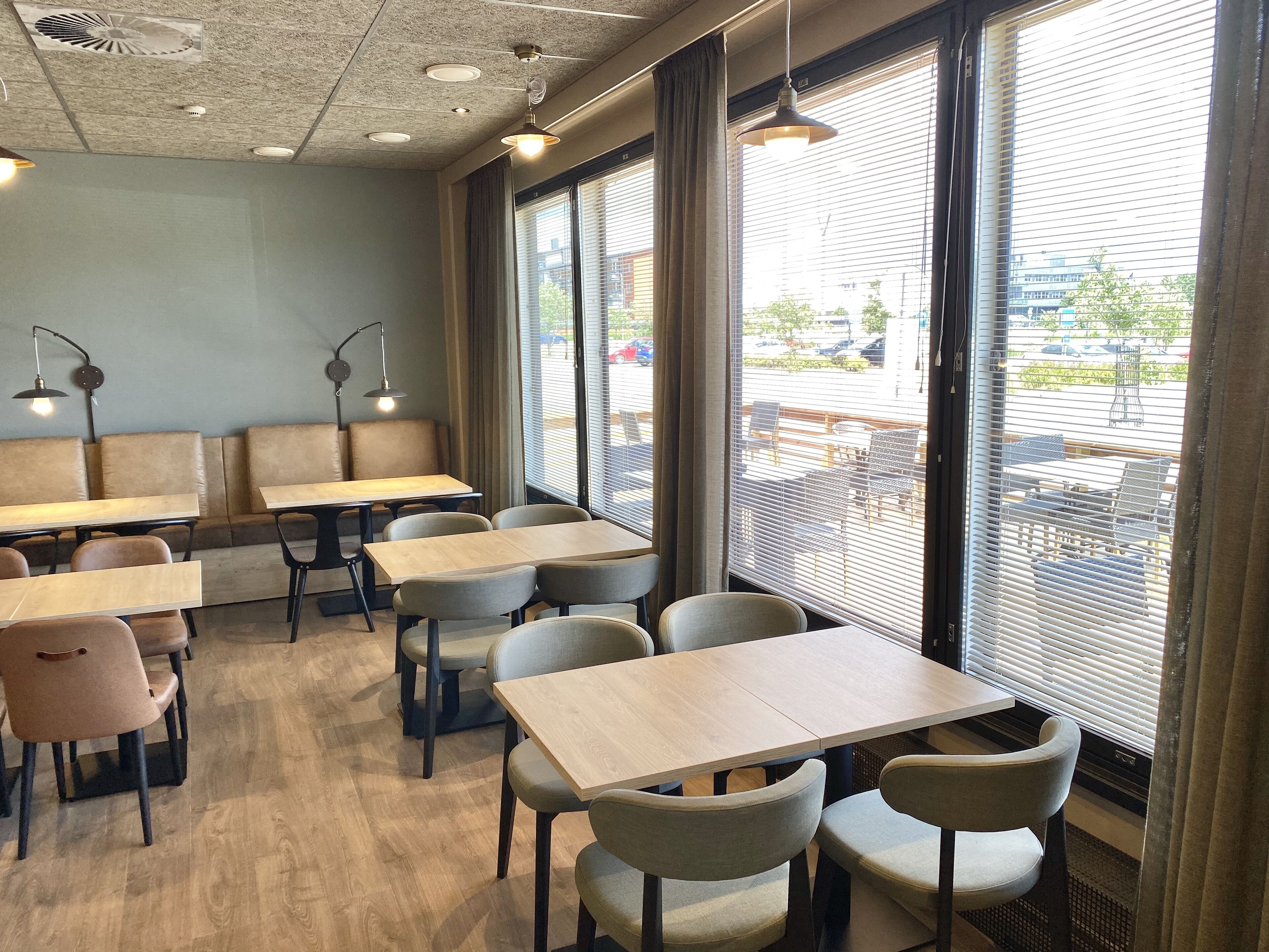 Un café moderno con mesas y sillas de madera, una zona de estar con bancos y grandes ventanales con persianas.