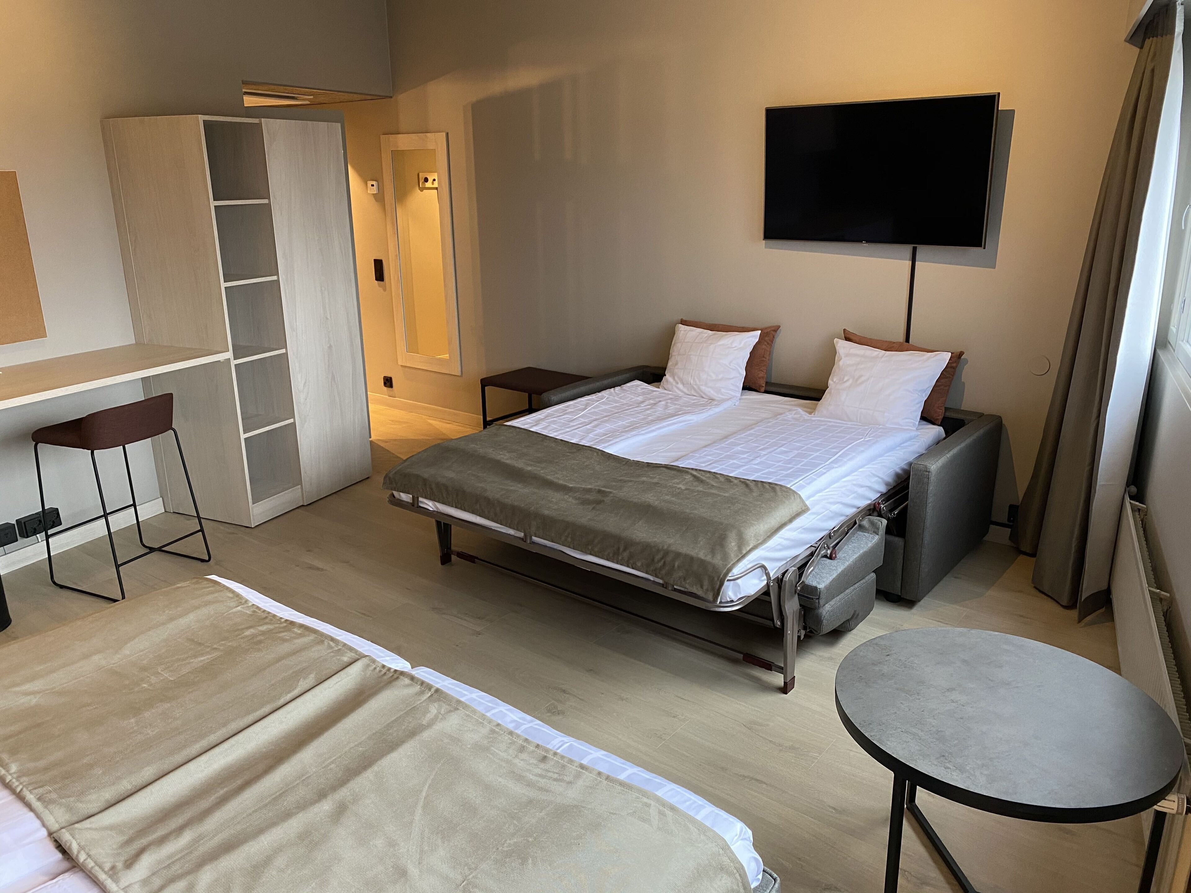 A hotel room with a bed, a desk, a chair, a TV, and a small table.