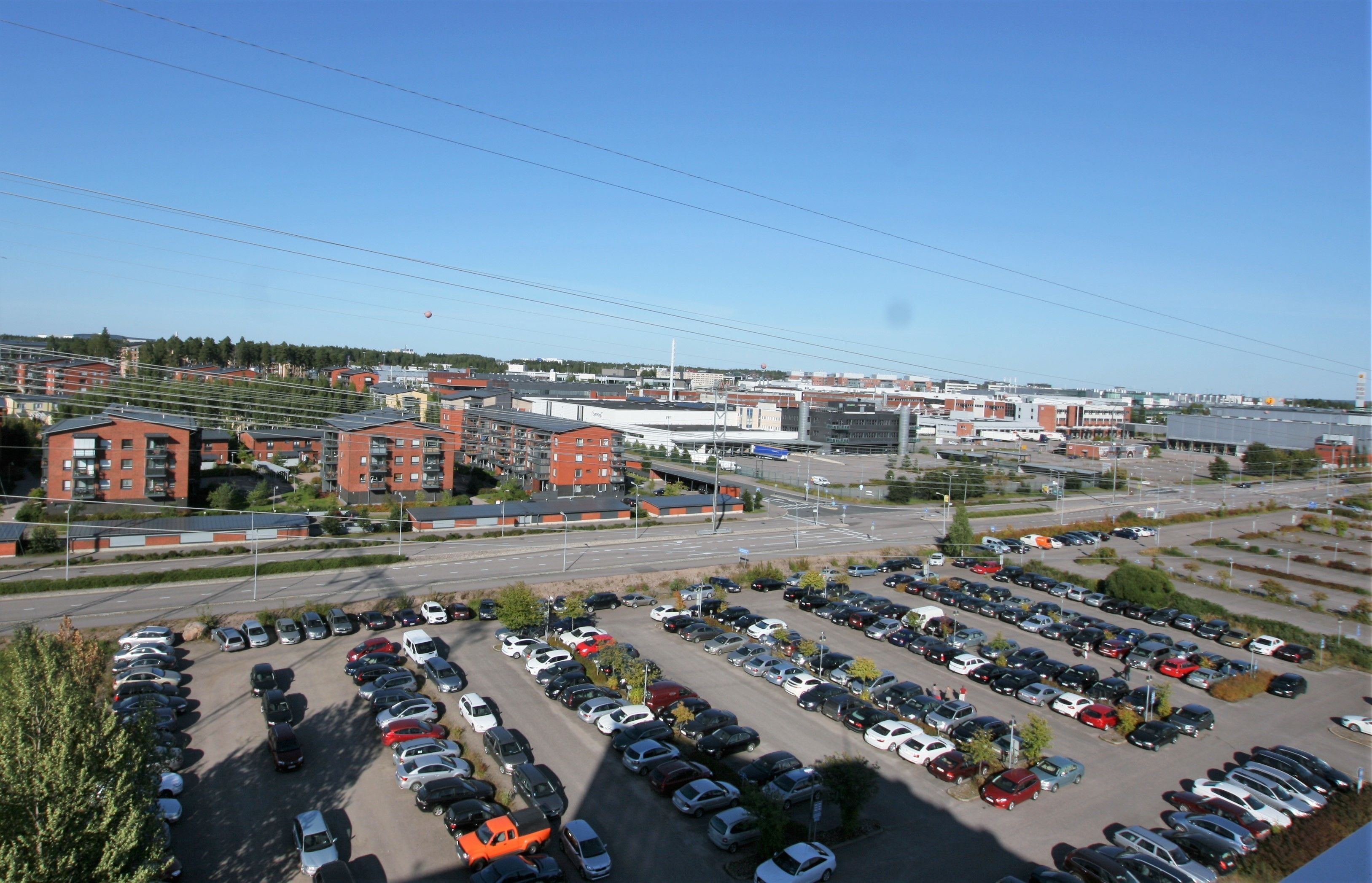 A parking lot with multiple rows of cars, a road with vehicles, and buildings in the background.