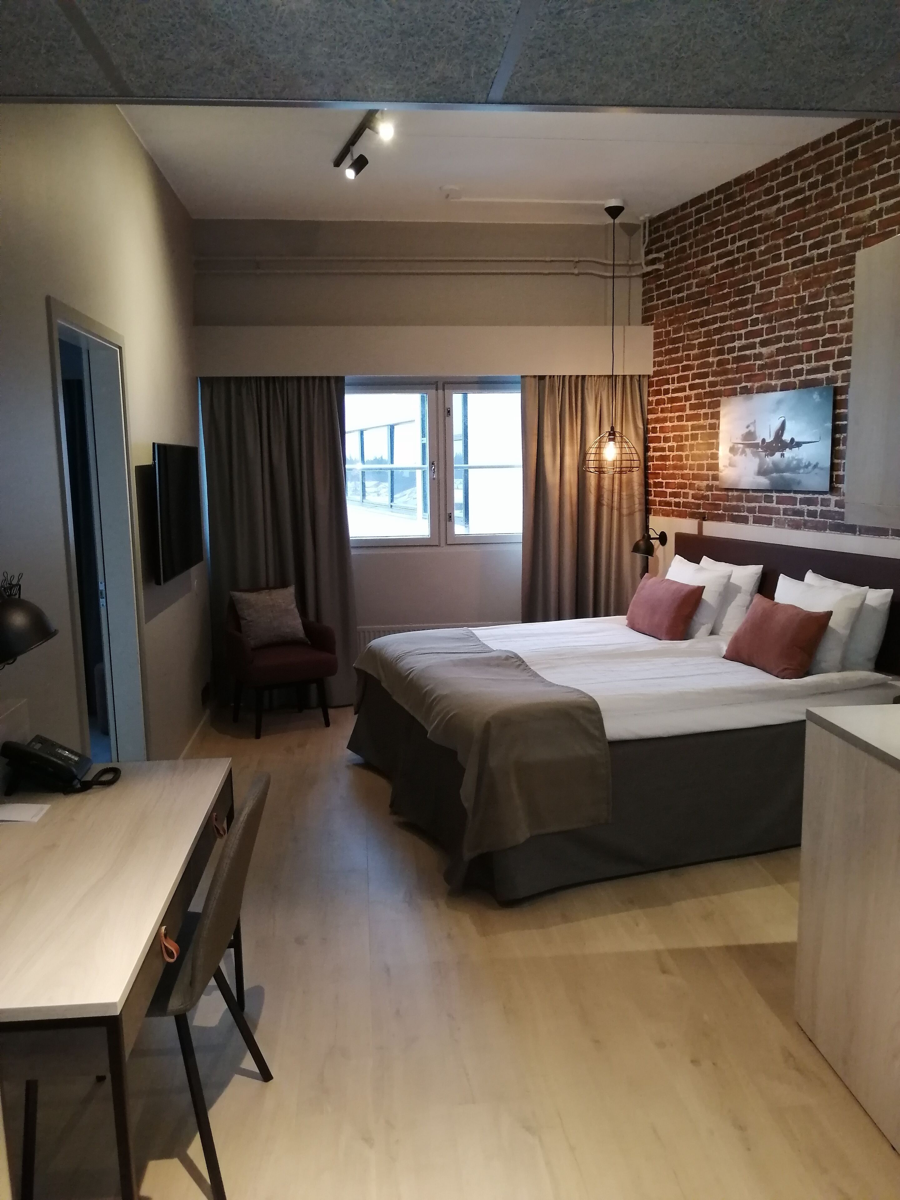 Moderni hotellihuone, jossa on sänky, työpöytä, tuoli ja ikkunasta avautuva näkymä ulos.