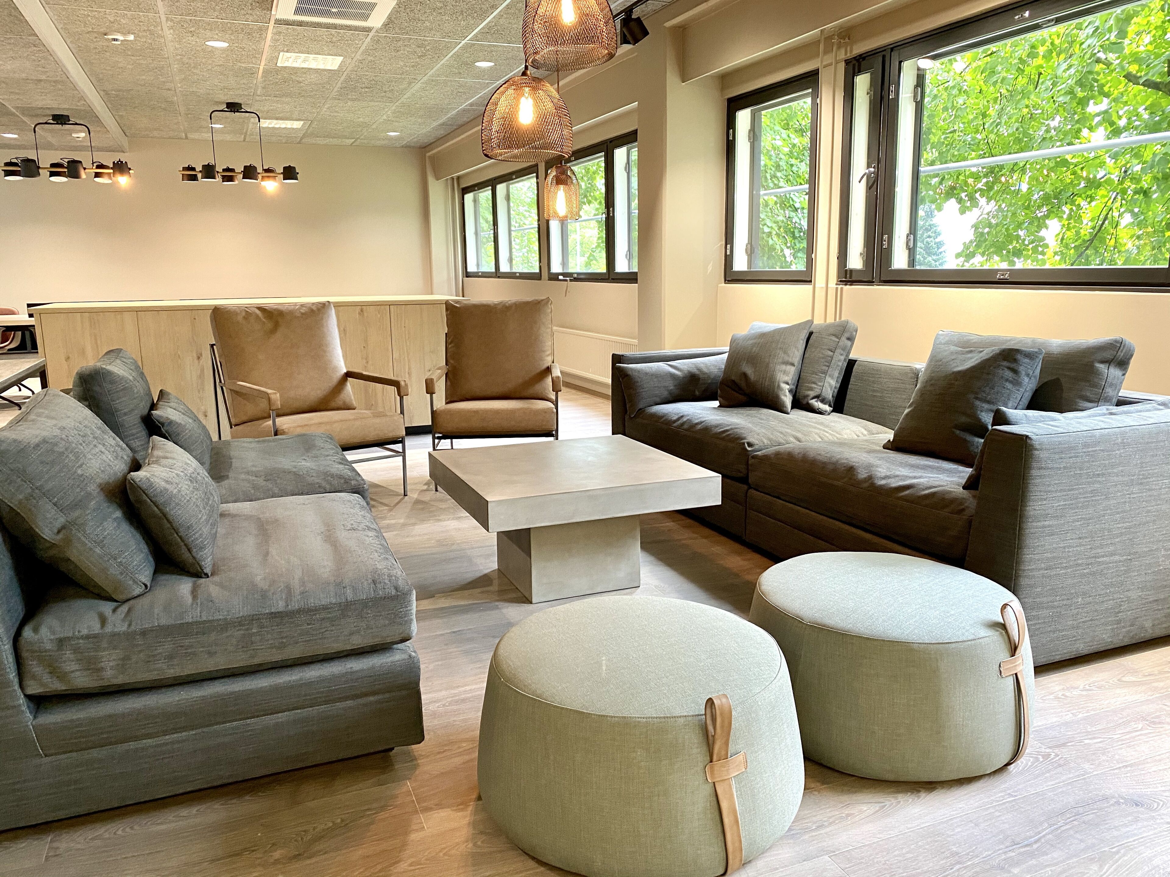 A modern living room with a sectional sofa, two armchairs, a coffee table, and ottomans. Large windows provide natural light and a view of greenery outside.