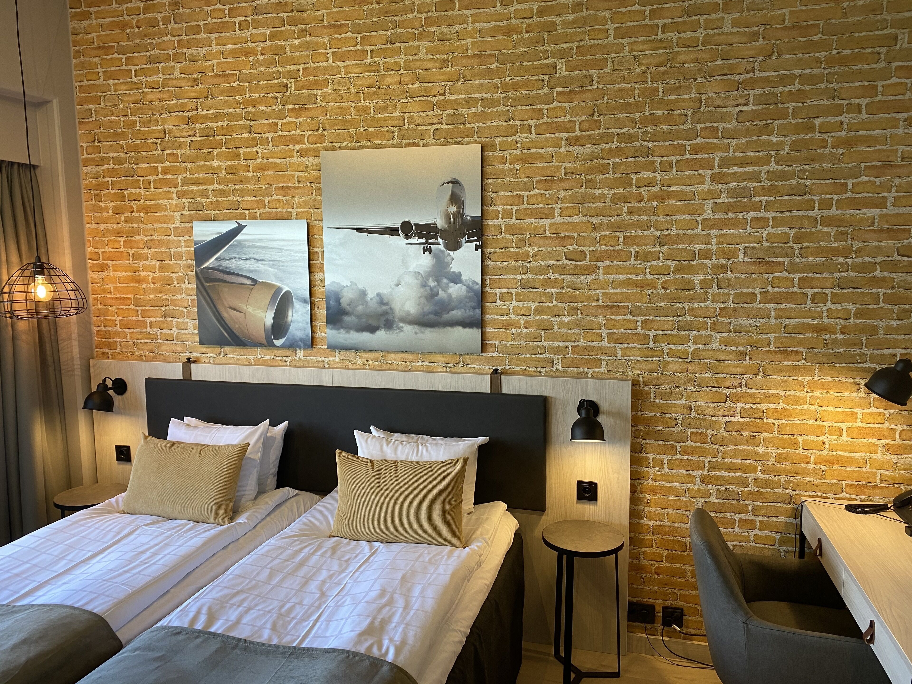 A hotel room with a brick wall, a large bed with white bedding, two bedside tables with lamps, and two wall-mounted artworks depicting an airplane and clouds.