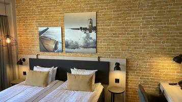 A hotel room with a brick wall, a large bed with white bedding, two bedside tables with lamps, and two wall-mounted artworks depicting an airplane and clouds.