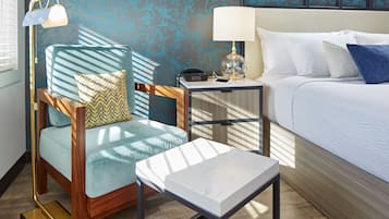 A hotel room with a bed, a desk with a lamp, a chair with a cushion, and a small table.