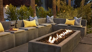 An outdoor seating area with a built-in fire feature, cushioned benches, and a small table with a bottle of wine and grapes.
