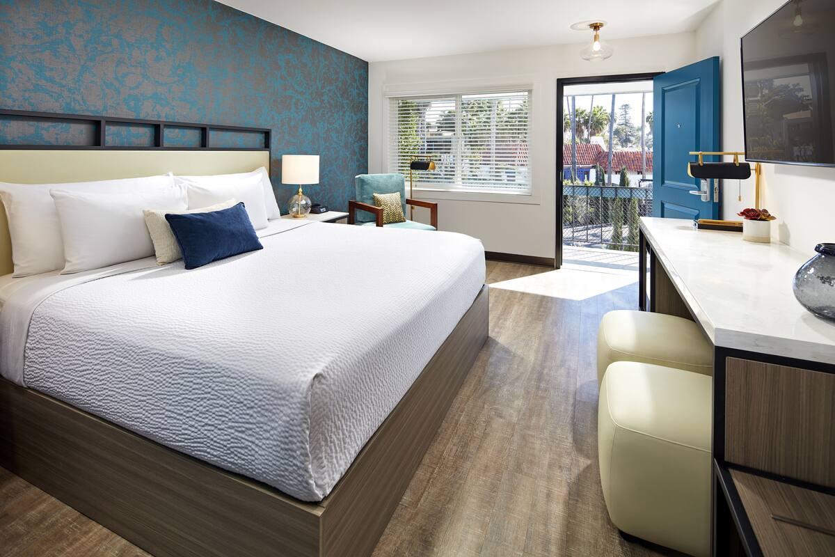 A hotel room with a large bed, a desk, and a chair. There is a window with blinds and a view of palm trees.