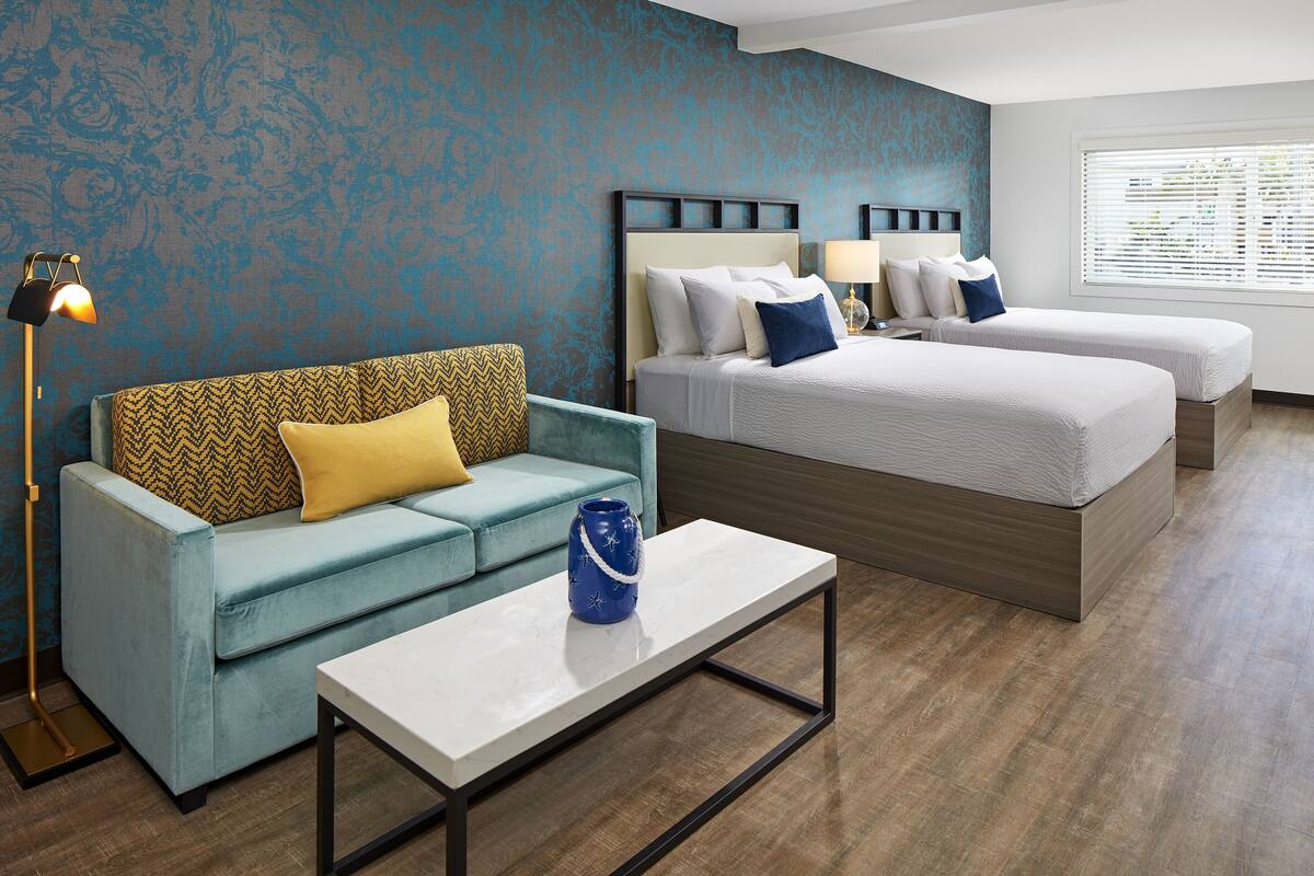 A hotel room with a blue patterned wall, a sofa with yellow and blue cushions, two beds with white bedding, a small table, and a floor lamp.