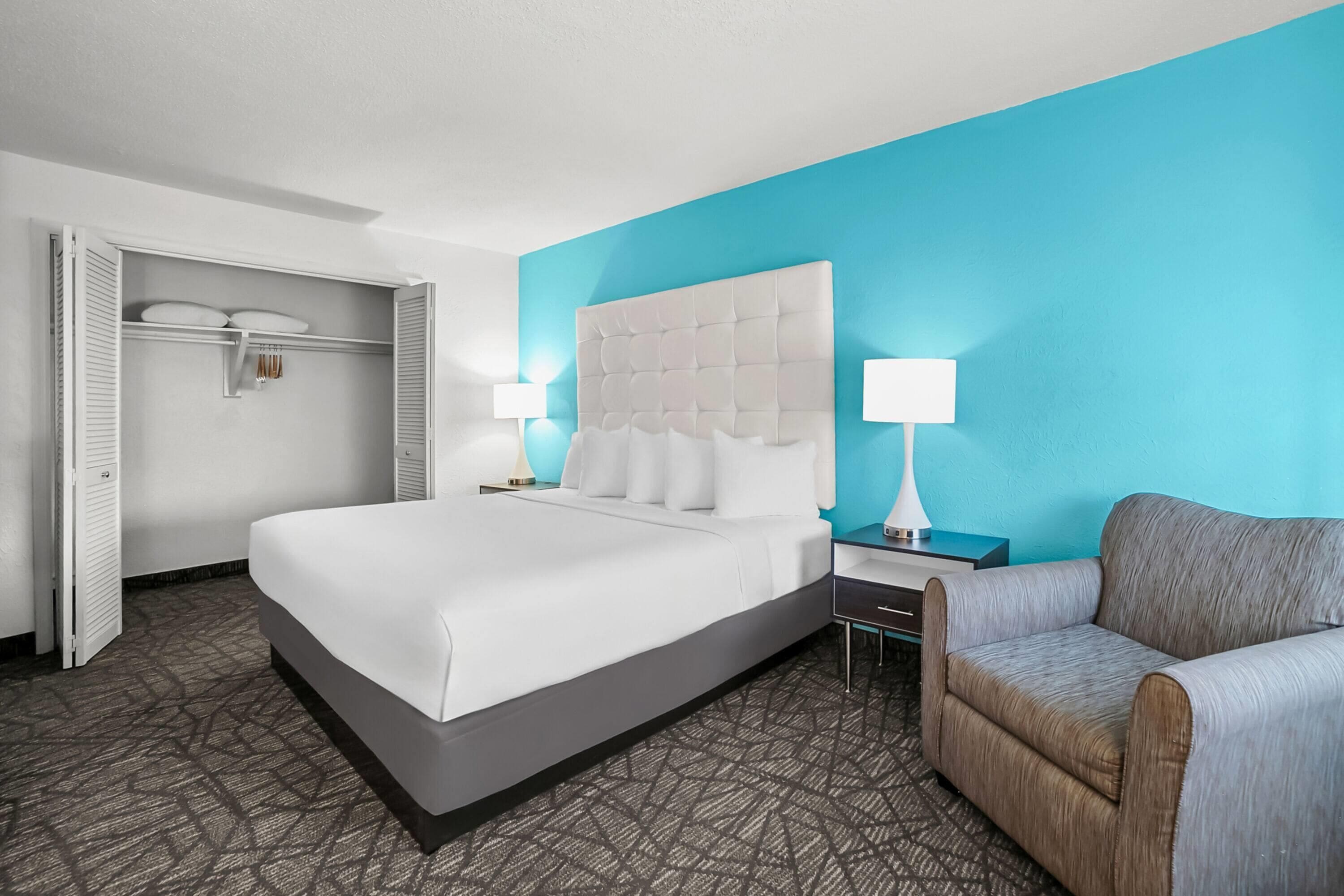 A hotel room with a large bed, a gray sofa, a nightstand with a lamp, and a blue accent wall.