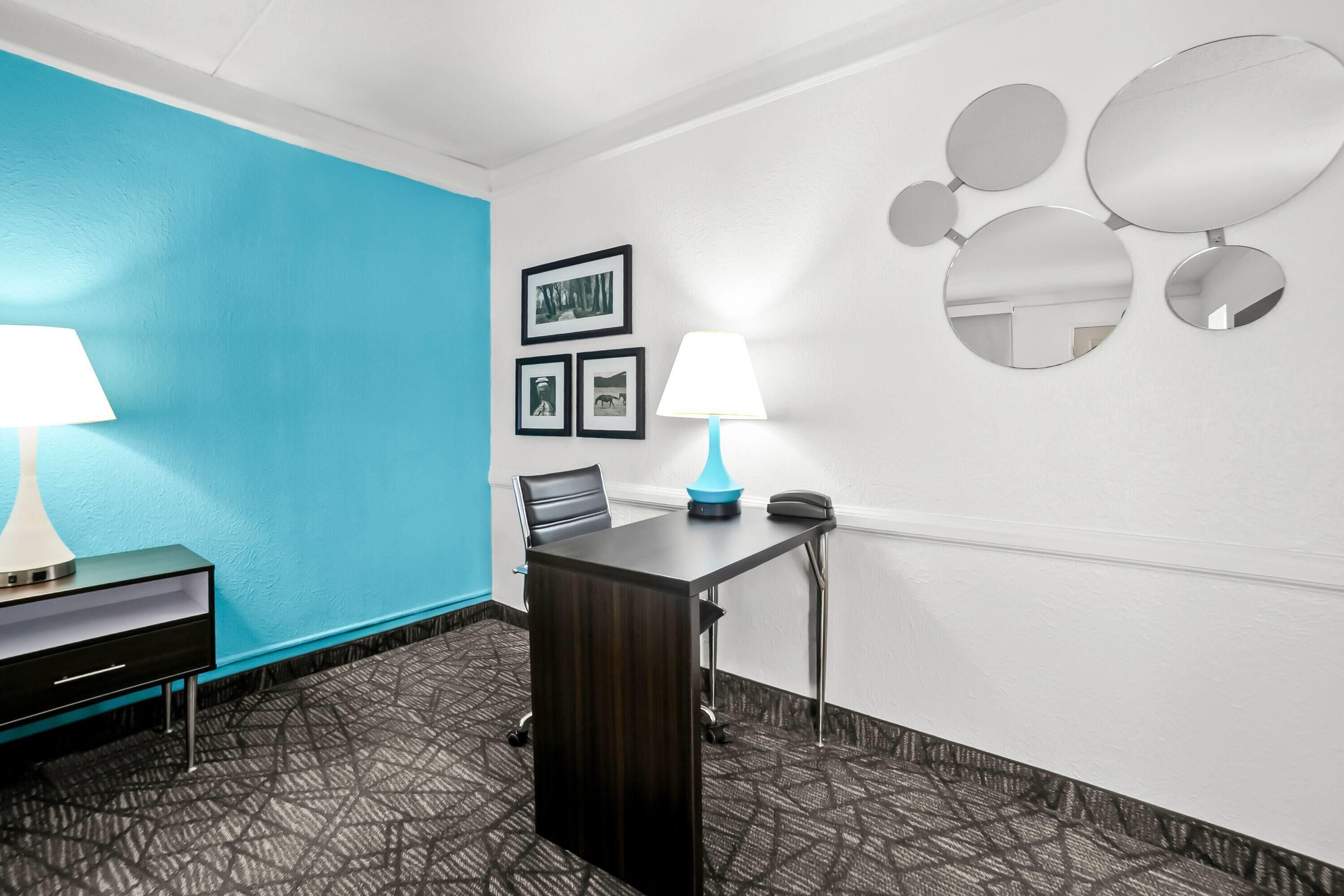 A hotel room with a dark wooden desk, a blue lamp, a chair, and a wall with framed pictures and mirrors.