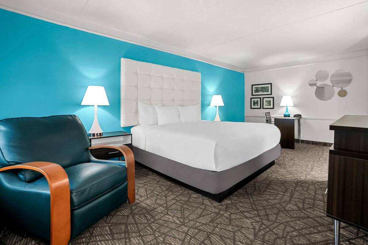 A hotel room with a large bed, a teal armchair, two bedside tables with lamps, and a dresser.