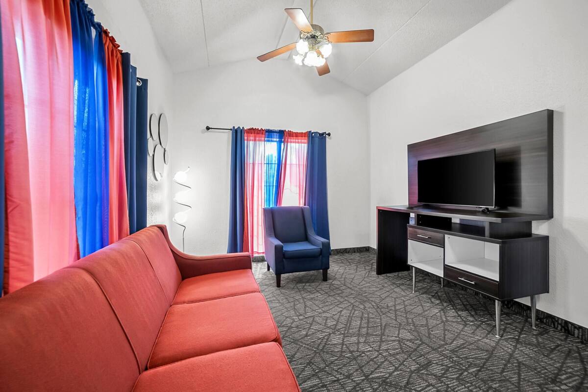 A hotel room with a red sofa, a blue armchair, a TV on a stand, and a ceiling fan.