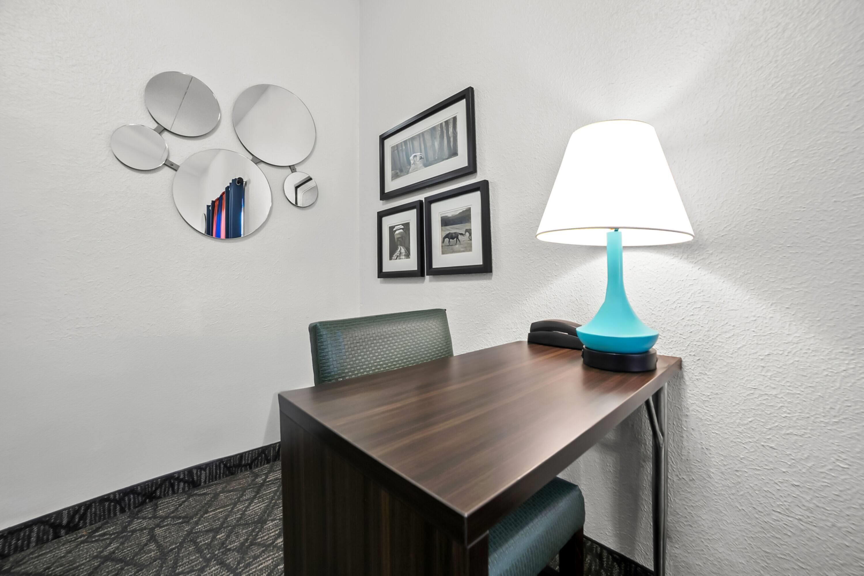 A hotel room with a wooden table, a blue lamp, a chair, and wall-mounted mirrors and framed pictures.