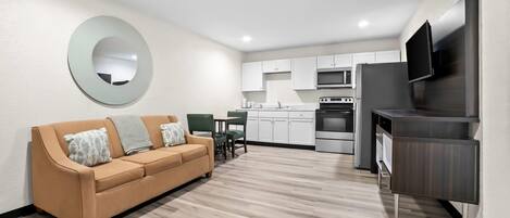 A modern living room with a sofa, a dining area, a kitchen with white cabinets, and a flat-screen TV mounted on the wall.