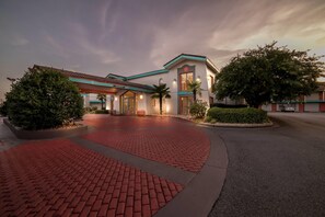 A hotel entrance with a red brick pathway leading to the main building, which features a modern design with a flat roof and large windows. - Howard Johnson by Wyndham Columbus Fort Benning (Columbus)
