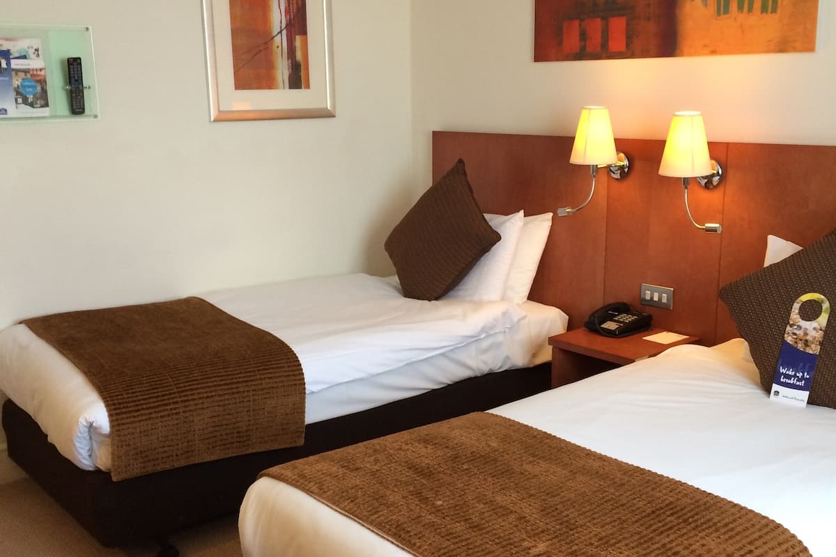 A hotel room with two single beds, each with a brown blanket and white linens. There are two wall-mounted lamps on the headboard, a framed picture on the wall, and a small table with a phone and a sign.