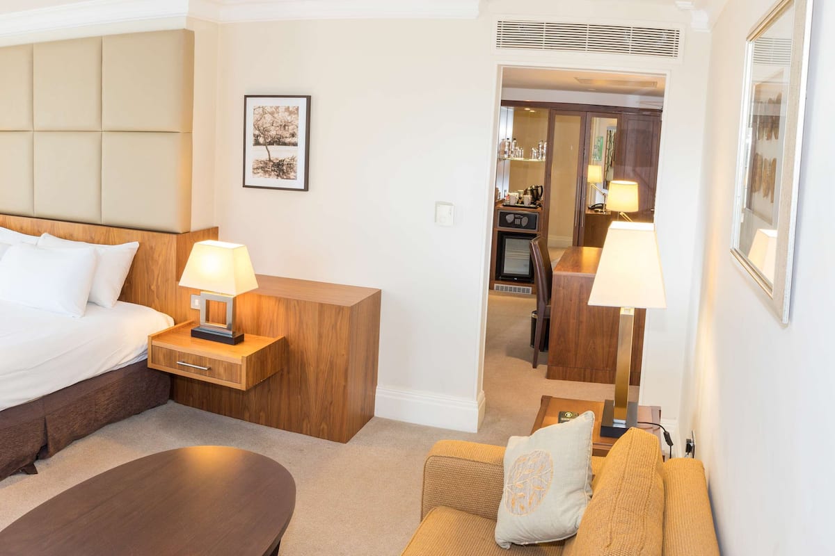 A hotel room with a large bed, bedside tables with lamps, a wooden headboard, a small round table, a sofa with cushions, and a view into the kitchen area.