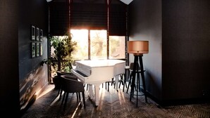 A modern room with a piano, a dining table, chairs, a lamp, and a large window with blinds. - Best Western Plus Moat House Reading (Wokingham)