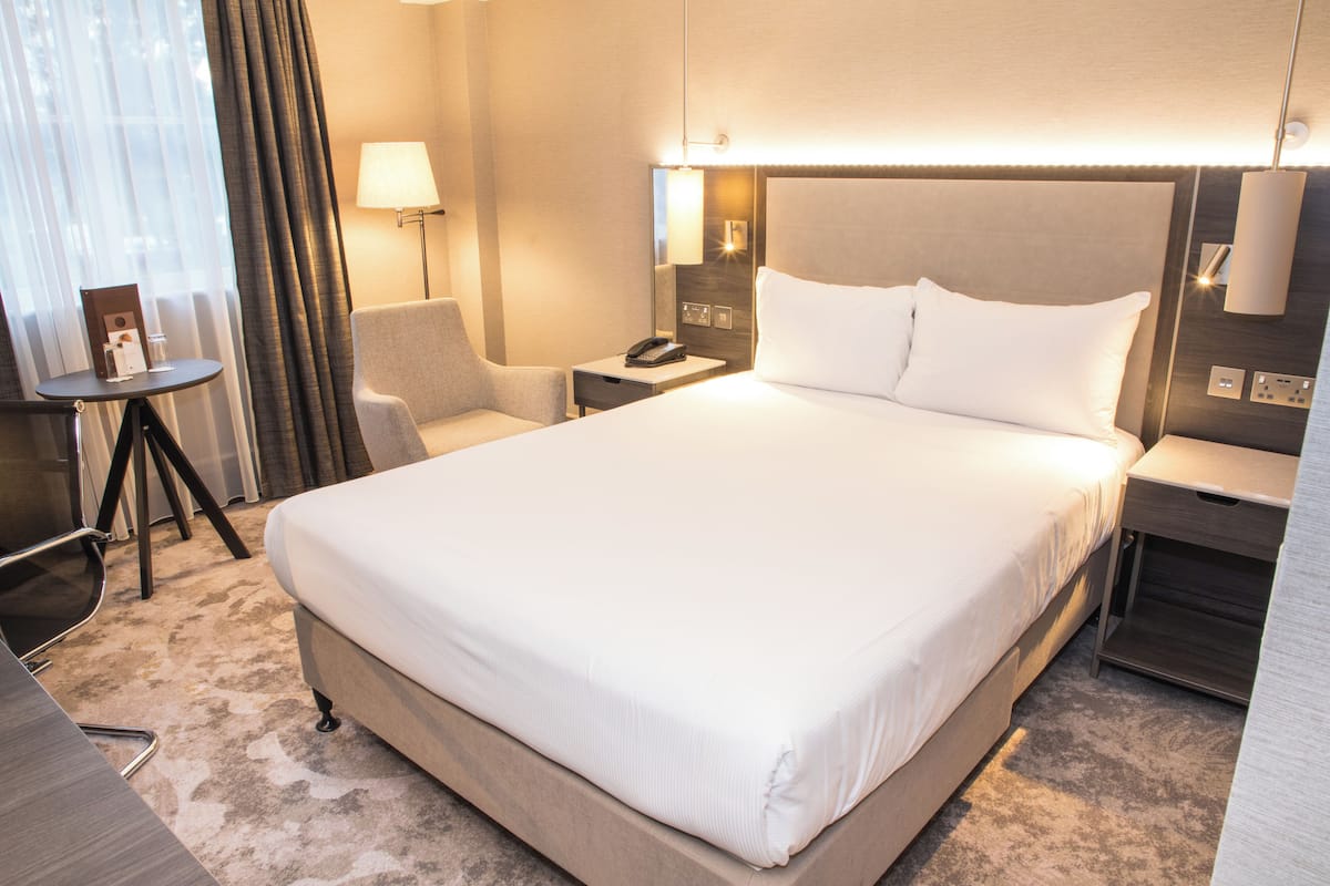 A hotel room with a large bed, a bedside table with a lamp, a chair, and a small table with a phone.