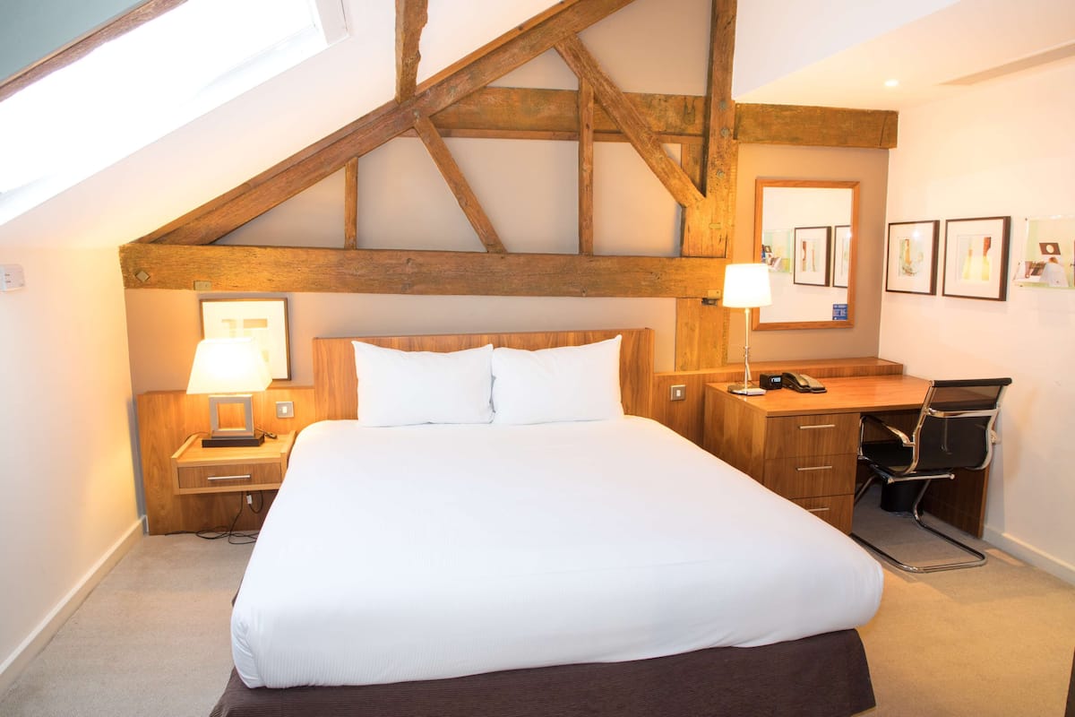 A hotel room with a large bed, wooden headboard, a desk with a chair, and a desk lamp.