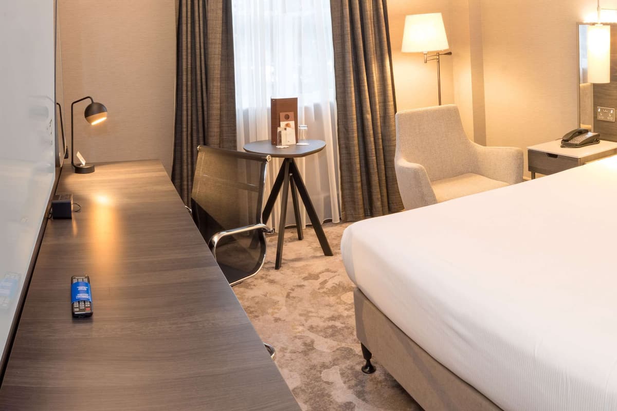 A hotel room with a large bed, a desk, a chair, a small table, and a lamp.