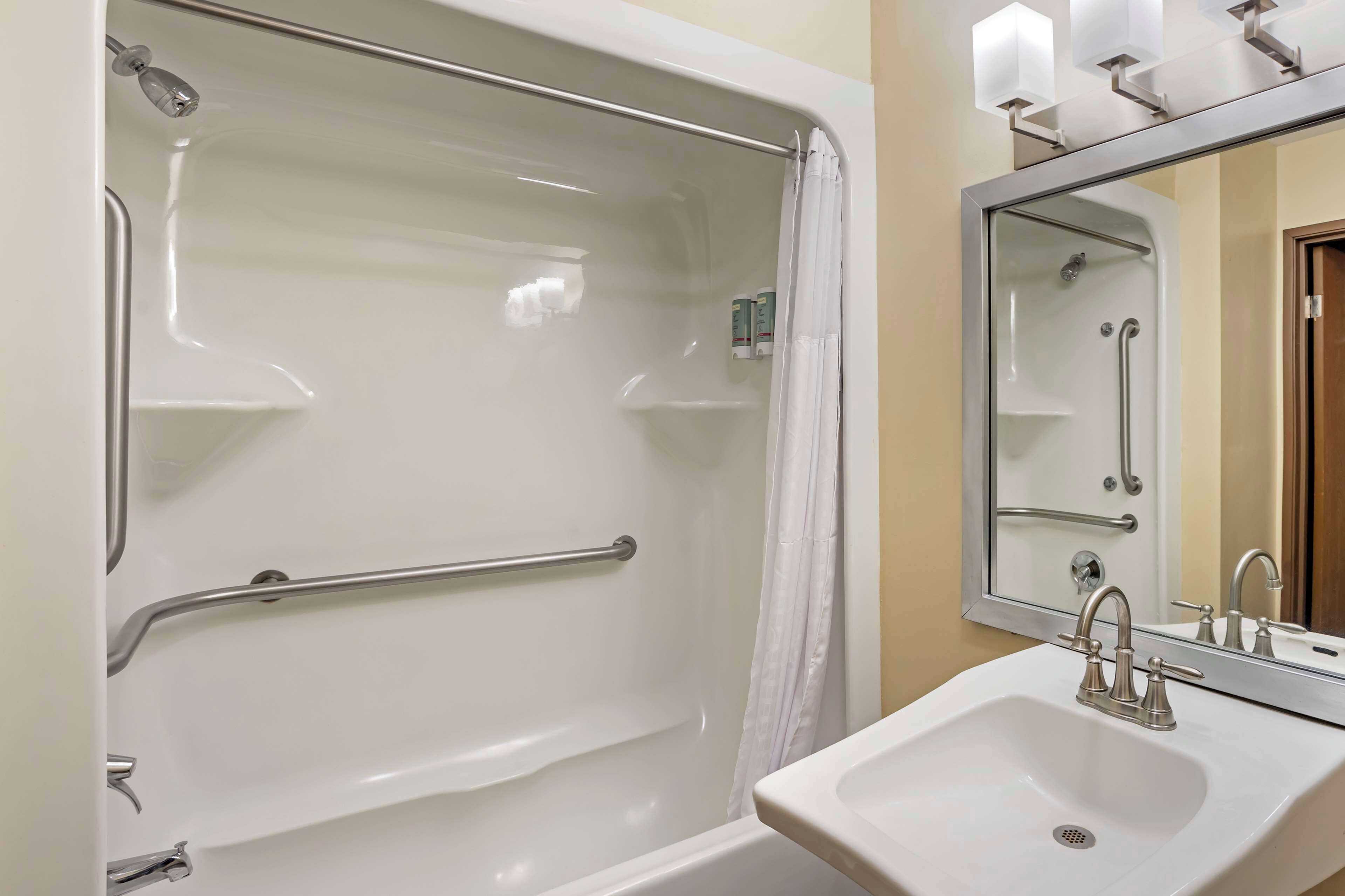 A bathroom with a white bathtub, a handrail, a shower curtain, a sink with a mirror, and a faucet.