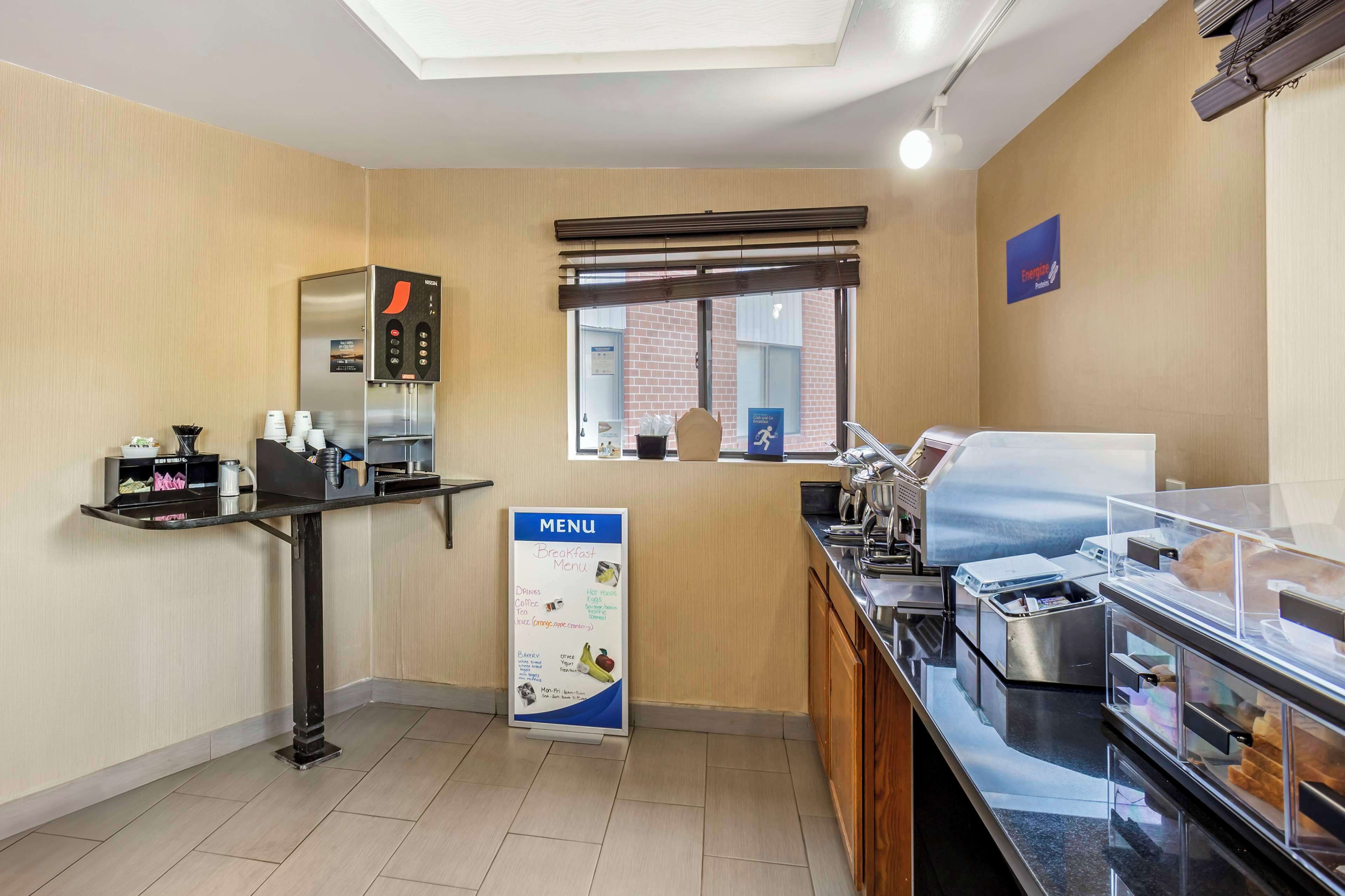 A hotel breakfast area with a self-service station, a menu board, and a window with blinds.