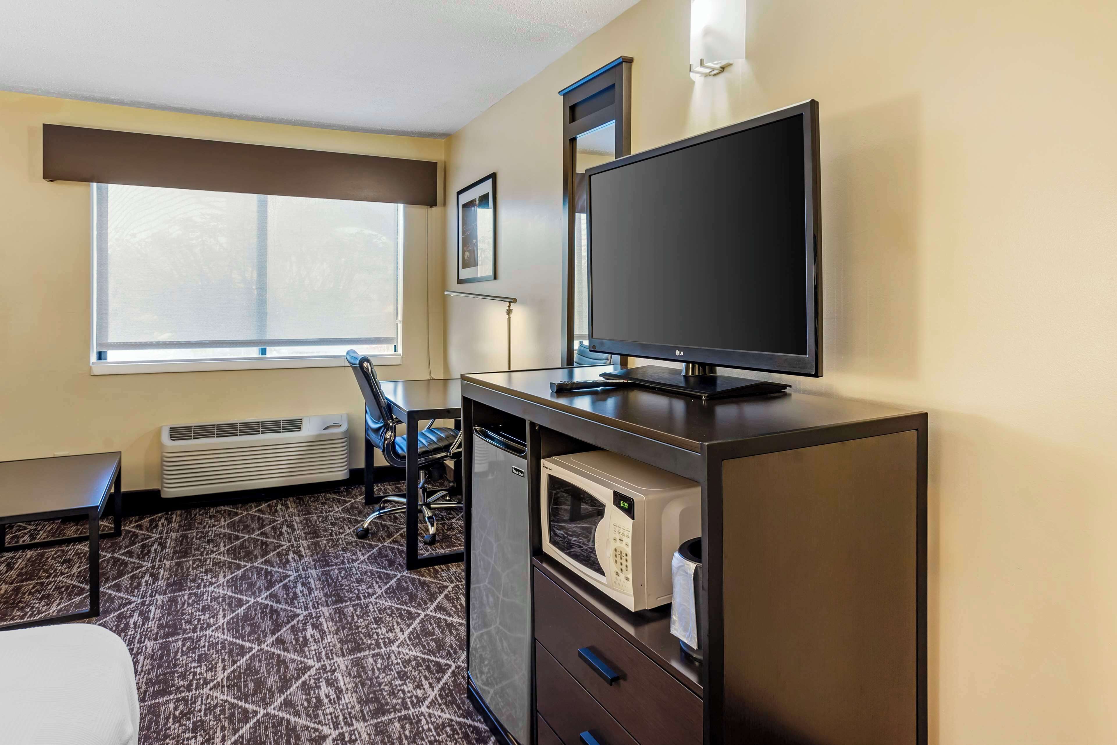 A hotel room with a flat-screen TV, a microwave, a desk, a chair, and a window with blinds.