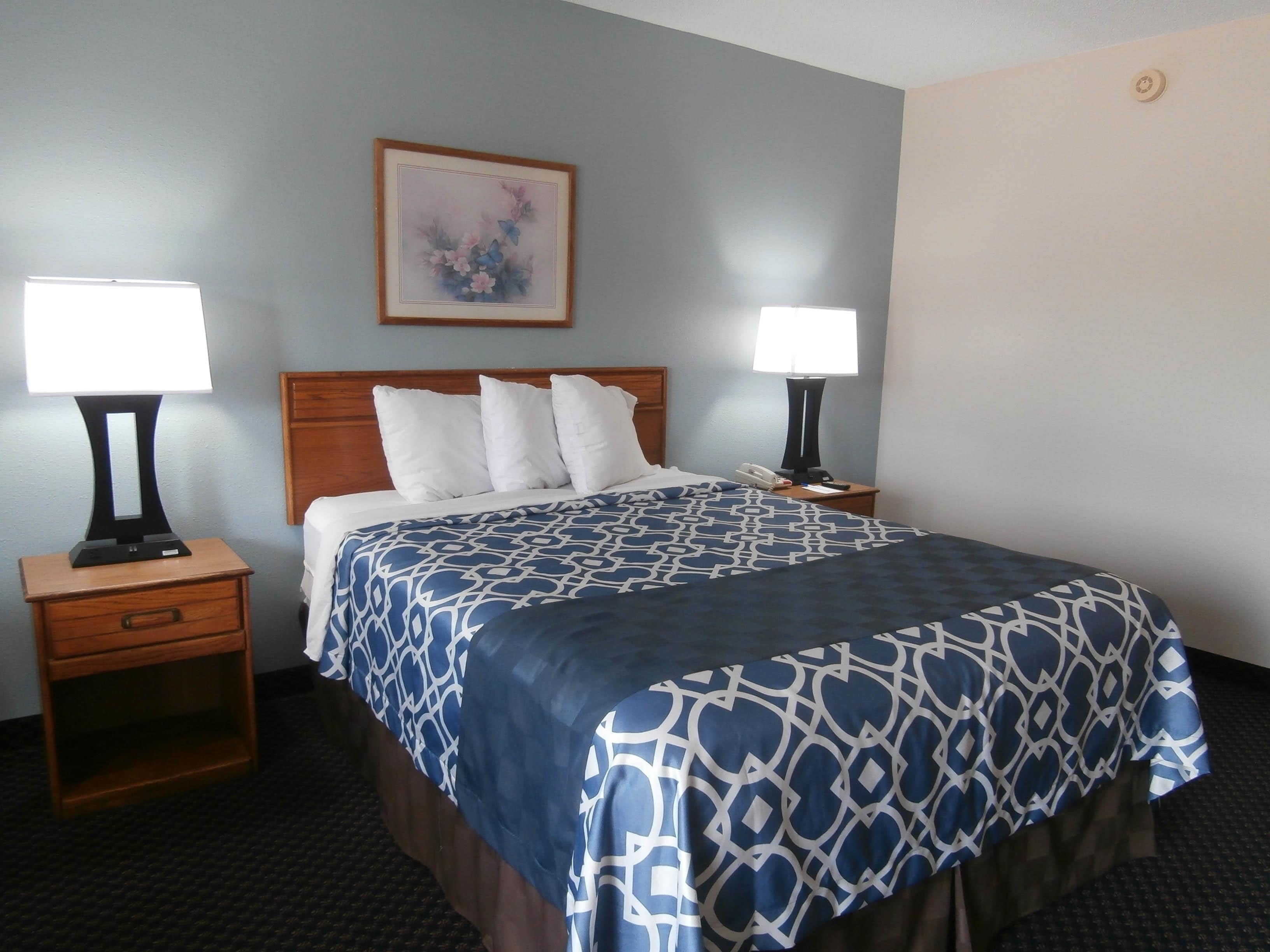 A hotel room with a bed, two bedside tables with lamps, a nightstand with a drawer, a framed picture on the wall, and a carpeted floor.