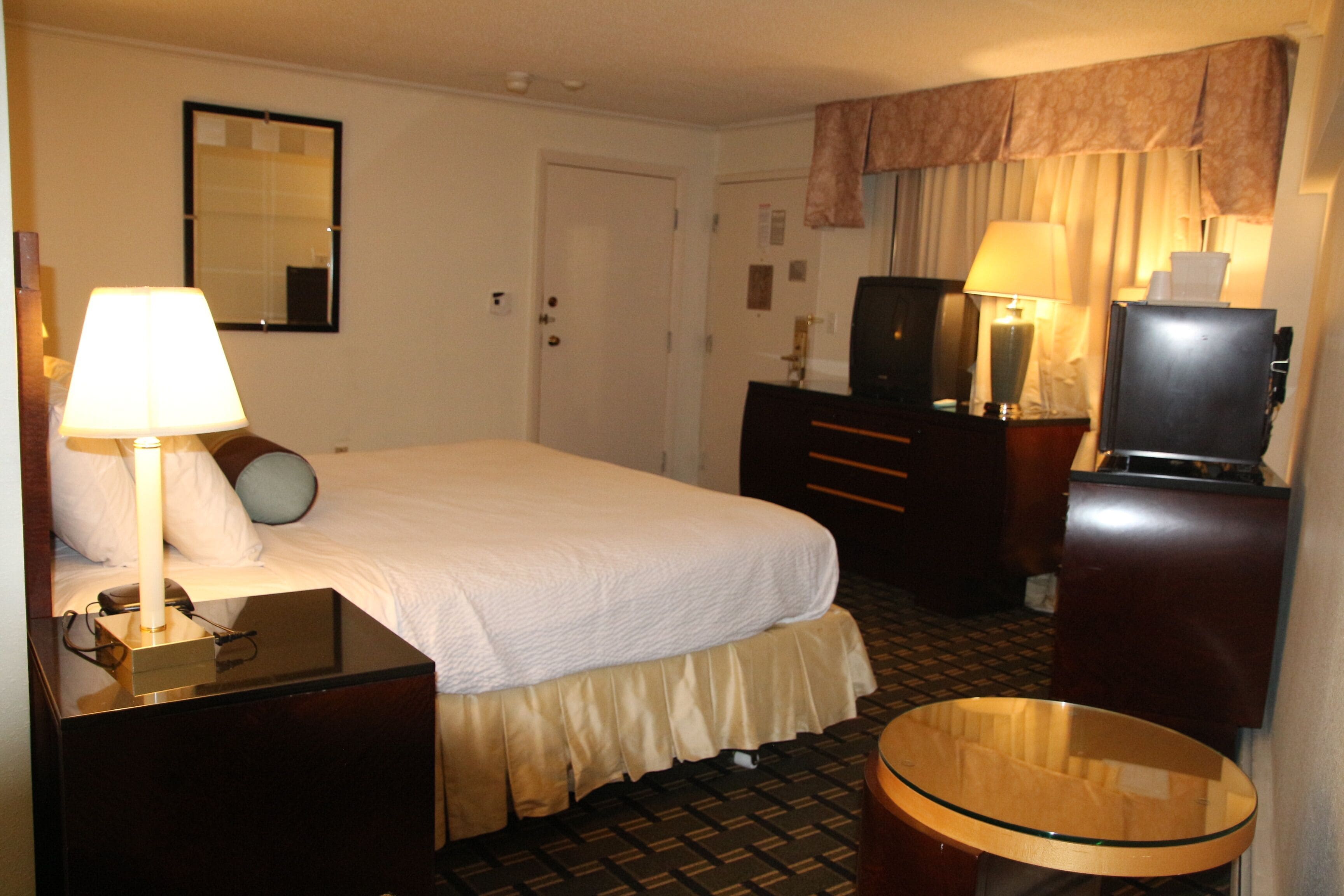 A hotel room with a bed, bedside table, lamp, dresser, mirror, and a small round table.