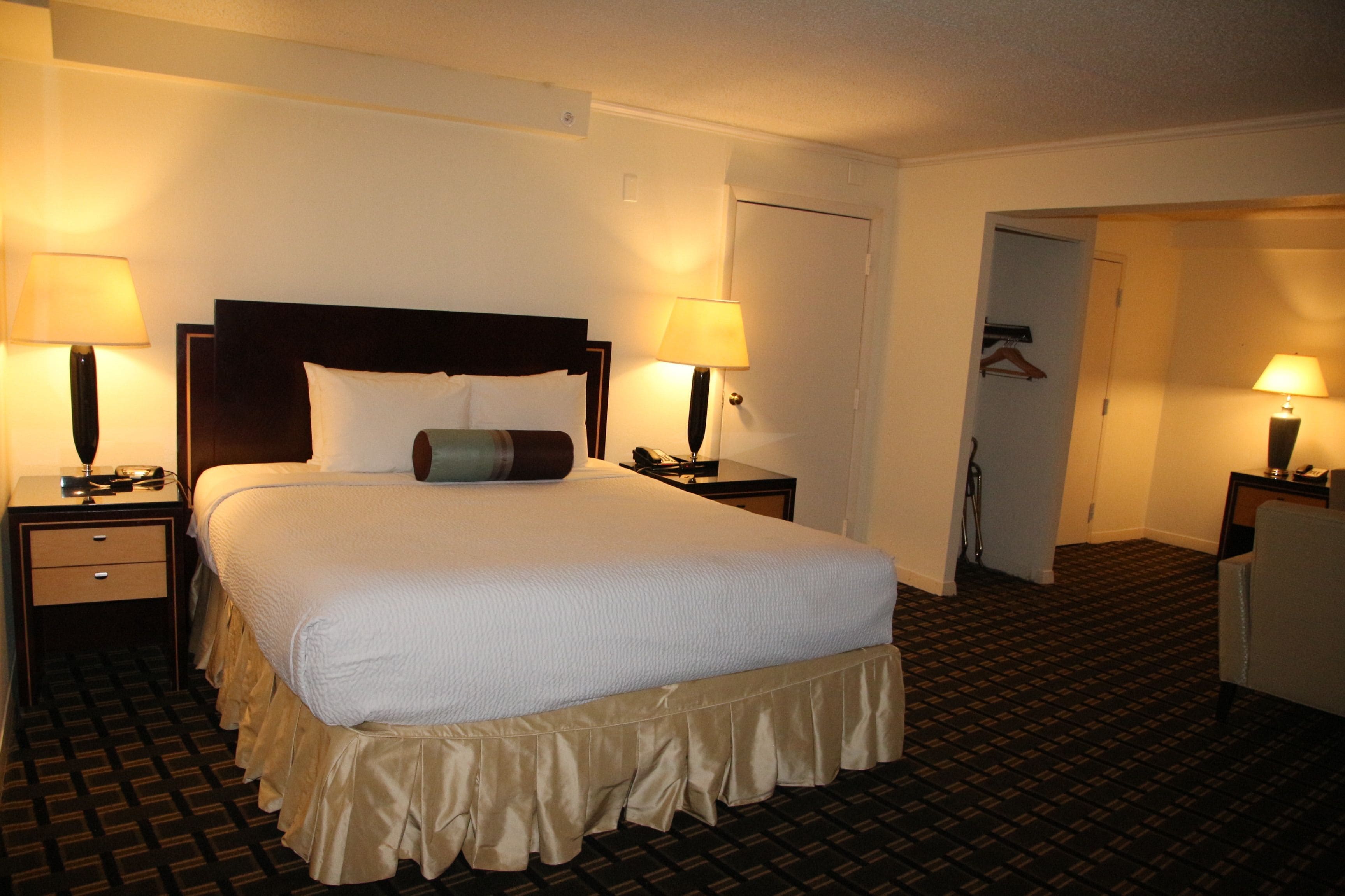 A hotel room with a large bed, two bedside tables with lamps, a wardrobe, and a door leading to another room.