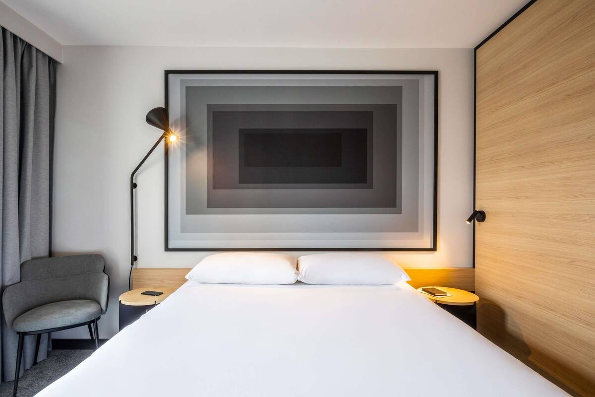 A hotel room with a large bed, a chair, a desk, a lamp, and a framed artwork on the wall.