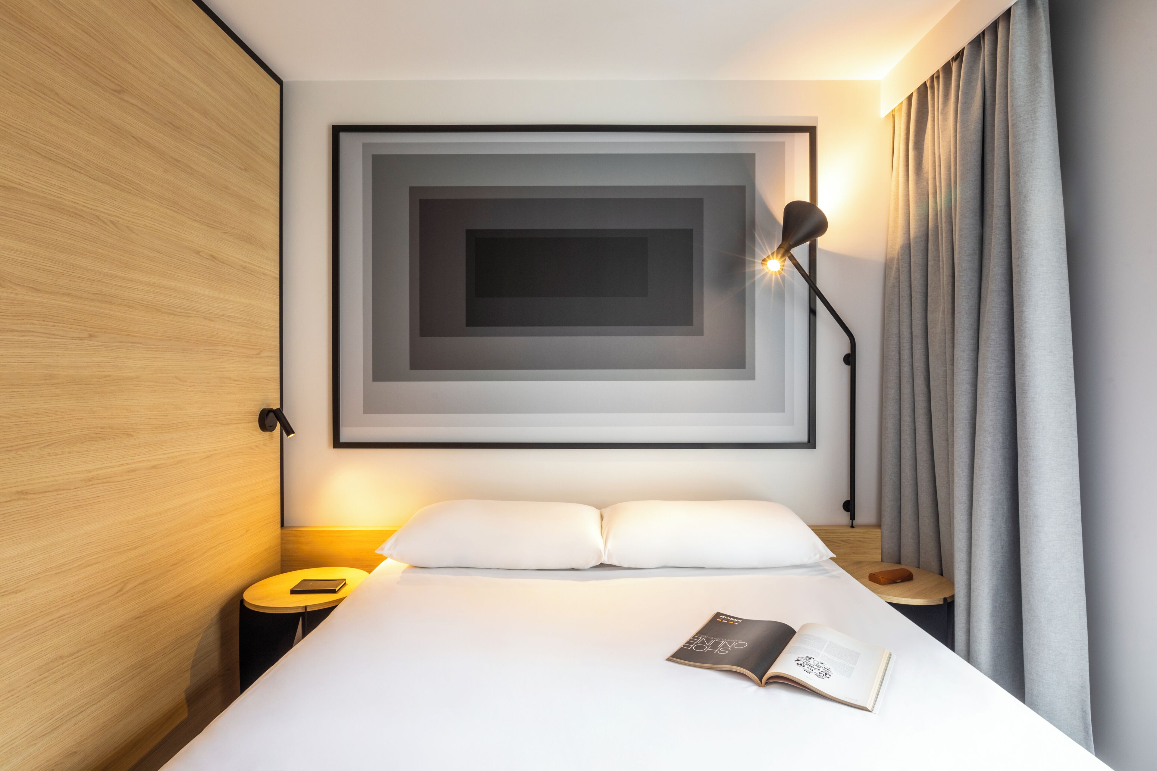 A modern hotel room with a neatly made bed, a bedside table with a lamp, a framed artwork above the bed, and a window with curtains.